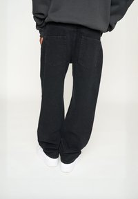 Person iført løse sorte jeans og hvide sneakers, der står mod en lys baggrund, set fra ryggen.