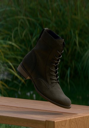 Σκούρο καφέ σουέντ μποτάκι με κορδόνια που αιωρείται πάνω από ένα ξύλινο τραπέζι με πράσινα φύλλα στο φόντο.