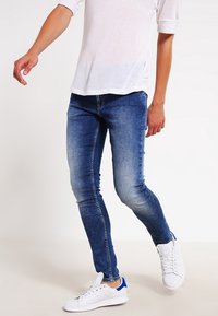 Slim fit blå denimjeans med en blekt utseende och subtila veckdetaljer. Kombinerad med vita sneakers. Enkel design.