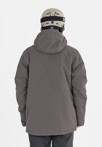 Grå skijakke fremstillet af slidstærkt stof, med hætte, lange ærmer og et enkelt design. Kombineret med en beige hjelm.