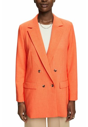 Kobieta ubrana w dwurzędowy, jaskrawo pomarańczowy żakiet z ciemnymi guzikami, biały top, beżowe spodnie oraz srebrny łańcuszek na szyi i bransoletkę.