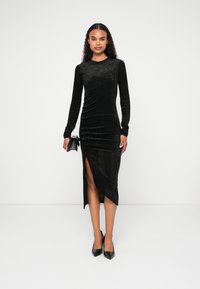 Rochie lungă din catifea neagră cu mâneci lungi și o fanta laterală. Prezintă o siluetă ajustată și un finisaj texturat. Asortată cu pantofi cu toc stiletto negri.