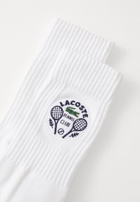 Bílé bavlněné ponožky s krátkým střihem a ribbenou texturou. Vyšívané kulaté logo s nápisem "LACOSTE TENNIS CLUB," tenisovými raketami a zeleným krokodýlem.