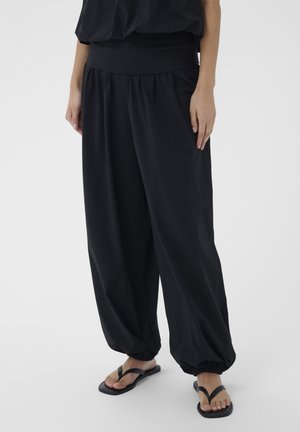 Személy laza fekete nadrágot visel, melynek szegélye összegyűrt, valamint fekete papucsot, és egy egyszerű, világos háttér előtt áll.