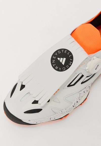 Vit sneakers med orange detaljer, texturerad ovandel och en unik remdesign. Har en svart cirkulär logo och ett BOA snörsystem.