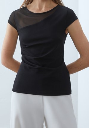 Vrouw draagt een strak zwart topje met een transparant diagonaal paneel over de borst en witte broek tegen een effen lichte achtergrond.