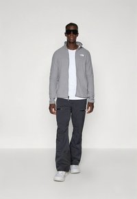 Gray zip-up hoodie, white t-shirt, and dark pants with zippers. Model wearing sunglasses; white shoes complete the outfit.