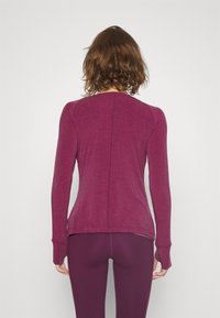 Langärmelig athleisure top in diep paars met een getailleerd ontwerp, naadloze schouders en verlengde manchetten, gecombineerd met bijpassende leggings.