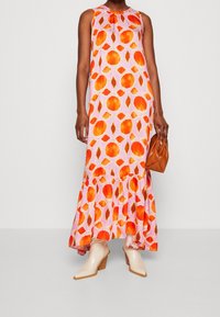 Ärmlös maxiklänning i rosa med orange och röda cirkulära mönster, med en volangkant. Kombinerad med beige ankelstövlar och en brun väska.