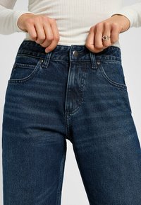 Tummansiniset farkut korkealla vyötäröllä, viiden taskun suunnittelu ja hillitty kulutus. Varustettu metallinapilla ja vetoketjulla.