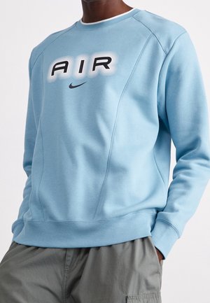 Mężczyzna w jasno niebieskiej bluzie z napisem "AIR" i logo Nike, w szarych spodniach bojówkach, ręce w kieszeniach.