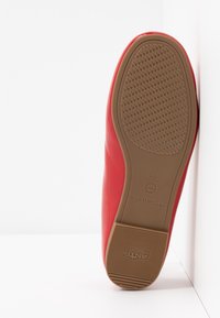 Chaussure rouge avec semelle antidérapante marron appuyée contre un mur blanc, montrant la texture de la bande de roulement et les marquages de taille 37.