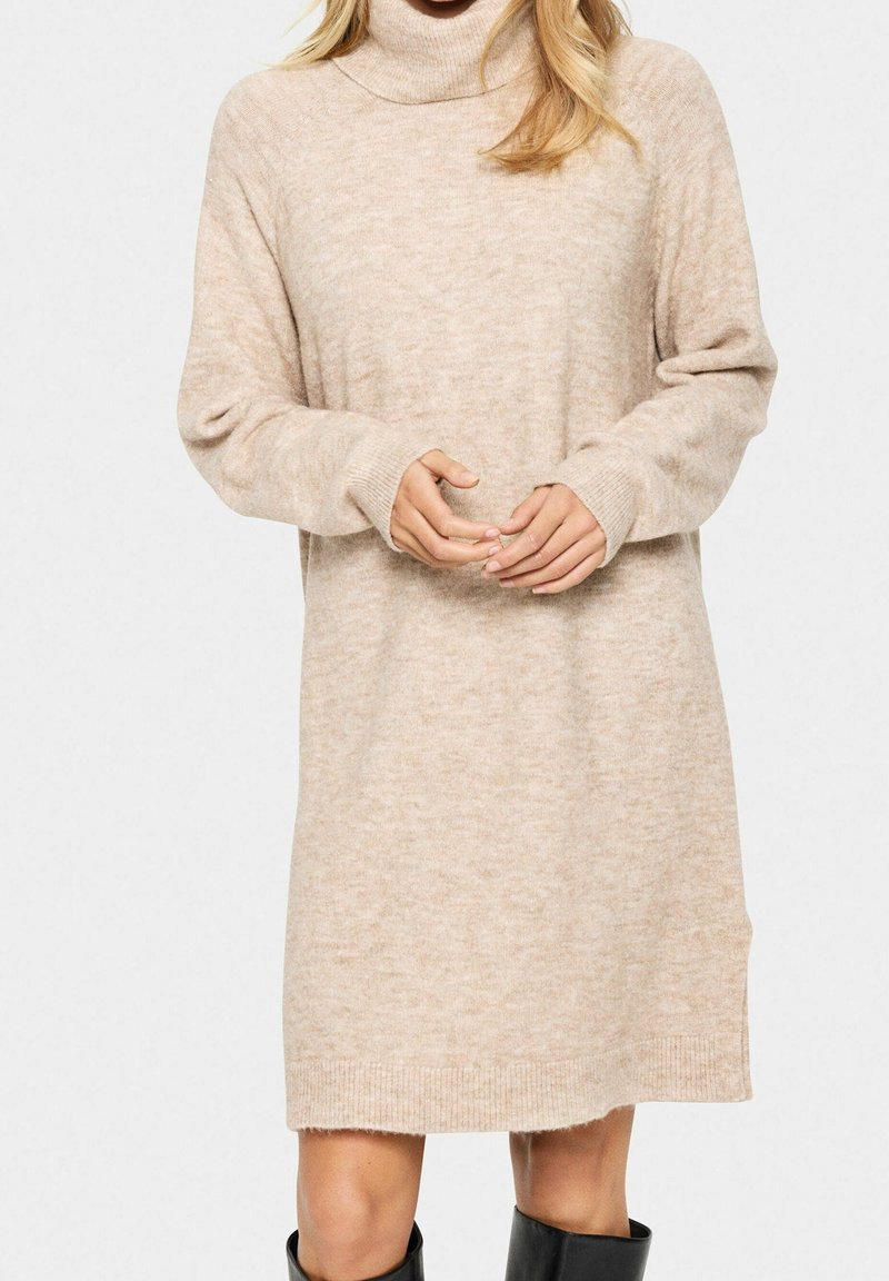 Femme portant une robe pull à col roulé beige en maille avec manches longues et bottes noires mi-mollet, debout devant un fond uni.