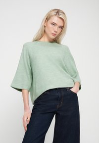 Pull surdimensionné vert menthe avec un col rond et de larges manches, associé à un jean bleu foncé à jambes larges. Texture douce et coupe décontractée.
