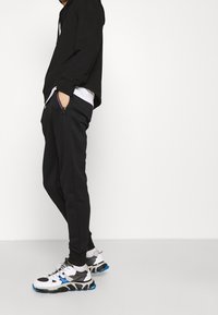 Schwarzer Hoodie mit lässiger Passform, kombiniert mit schwarzen schmal geschnittenen Hosen. Weiße und graue Sneakers mit blauen Akzenten und Netzdetails.