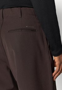 Brown pants with a textured finish, featuring a back pocket, logo detailing, and a straight cut design. Worn with a black long-sleeve top.