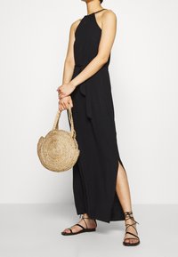 Czarna sukienka bez rękawów z dekoltem halter i bocznymi rozporkami. Trzyma okrągłą plecioną torbę. Posiada płaskie sandały na rzemykach. Gładka tekstura.
