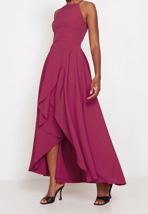 Vrouw draagt een mouwloze, hooggesloten fuchsia jurk met gelaagde ruches en zwarte open teen pumps op een witte achtergrond.
