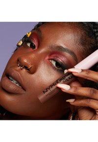 Matte flytande läppstift i mjuk brun förpackning med svart text. Modellen har djup brun hud, djärv ögonmakeup och flerfärgade naglar.