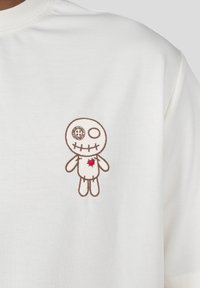 Camiseta de algodón blanca con un diseño bordado de muñeco vudú marrón que incluye un detalle de corazón rojo, colocado de manera destacada en el lado izquierdo.