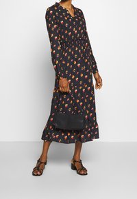 Musta pitkähihainen mekko kukkakuviolla, röyhelökaula-aukko, vyötäröltä kireä, levenevä helma, yhdistetty pieneen mustaan olkalaukkuun ja remmisandaaleihin.