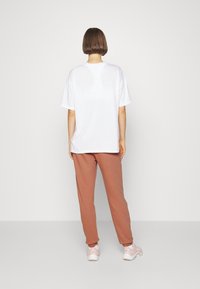 Valkoinen ylisuuri t-paita, lyhyet hihat, yhdistetty vaaleanruskeisiin verkkareihin, pehmeä kangas ja vaaleanpunaiset lenkkitossut, nähtynä takaa.