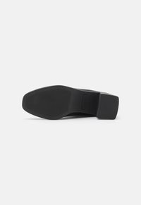 Semelle de chaussure noire, forme rectangulaire, motif de grip texturé, talon carré, finition lisse, design minimal, avec étiquette de marque emboutie.