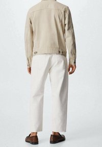 Chaqueta de mezclilla beige con corte recto, detalle de botones en la parte trasera, combinada con pantalones blancos y calzado marrón. Vista trasera, fondo neutro.