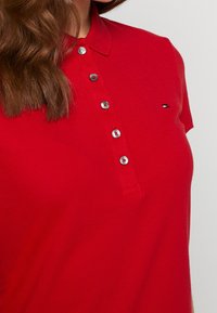 Czerwona koszulka polo wykonana z teksturowanej tkaniny, z klasycznym kołnierzykiem, krótkimi rękawami i srebrnymi guzikami. Mały akcent w postaci logo na piersi.