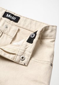 Pantalones de mezclilla color off-white con la cremallera parcialmente desabrochada, cierre de botón, etiqueta negra de la marca Molo en la cintura y botón de metal con el texto "Molo jeans".