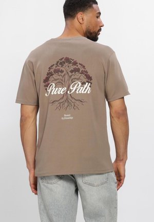 Man draagt een bruin T-shirt met een boomgrafiek en tekst "Pure Path" en "Rooted by friendship" op de rug, gecombineerd met lichtgrijze jeans.