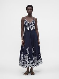 Femme portant une robe bleu marine sans manches avec une broderie florale blanche sur le corsage et l'ourlet, debout devant un fond clair uni.