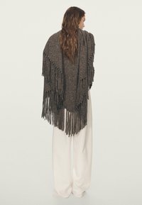 Poncho lavorato a maglia di colore marrone scuro con lunghe frange all'orlo. Indossato sopra pantaloni bianchi, presenta una superficie strutturata e un design aperto.