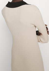 Abito maglia beige a coste con colletto nero, caratterizzato da maniche lunghe e una silhouette aderente, che mette in risalto la texture e il design slanciato.