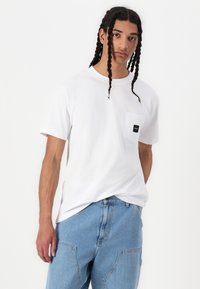 Tricou alb din bumbac, cu guler rotund și un buzunar pe pieptul stâng, având un eticheta neagră. Purtat cu pantaloni din denim albastru deschis.