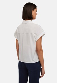 Blusa blanca de manga corta con cuello, que presenta un corte relajado y una textura suave; el diseño de la parte trasera incluye una costura central.