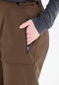 Bruine outdoorbroek met een zijrits zak, gemaakt van een gestructureerde, duurzame stof. Gedragen met een grijze langemouwen top.
