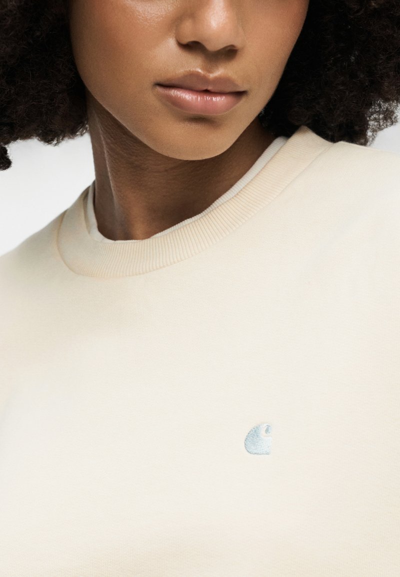 Hellbeiges Sweatshirt mit geripptem Rundhalsausschnitt und kleinem, blau gesticktem Logo auf der Brust. Das Material wirkt glatt und weich.