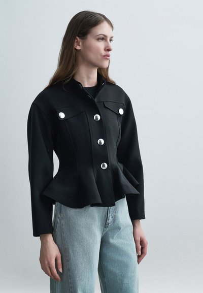 Femme portant une veste noire ajustée avec de gros boutons argentés et un jean bleu clair, debout devant un fond uni clair.