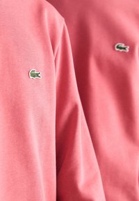 Sudadera rosa de algodón con cuello acanalado, que presenta un pequeño logo de cocodrilo verde en el pecho. Textura lisa con un ajuste relajado.