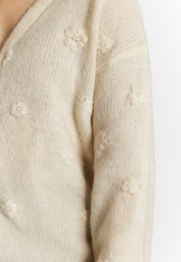 Beige stickad cardigan med V-ringning, prydd med broderade blommor över hela tyget och en mjuk, strukturerad yta.