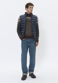 Navy Blousonweste über einem braunen gemusterten Pullover, lässige blaue Jeans und dunkelbraune Stiefel. Strukturierte Stoffe und kontrastierende Farben sind vorhanden.