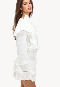 Witte jurk met gelaagde ruffles, gestructureerde stof, lange mouwen en subtiele verticale strepen. Opvallende franjes accent aan de onderrand.