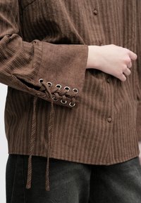 Ruda dryžuota marškinėliai su tekstūruota medžiaga, rankogalyje su nėriniais ir akių detalėmis. Priekyje esantys mygtukai suteikia funkcionalumo.