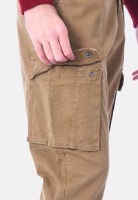Pantaloni cargo in cotone khaki con una tasca laterale, chiusa da bottoni a pressione; il tessuto appare resistente e leggermente testurizzato. Primo piano sulla zona della tasca.