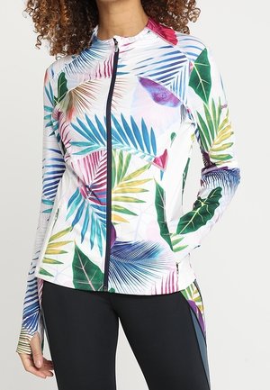 Frau trägt eine weiße Reißverschluss-Jacke mit bunten tropischen Blattmustern, kombiniert mit schwarzen Leggings, posiert mit einer Hand in der Tasche.