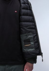 Svart långärmad tröja under en zippered pufferjacka. Jackan har en mjuk, blank svart yta och en grön ficka med en varumärkeslogo.