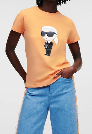 Persona que lleva una camiseta color melocotón con una figura de dibujo animado con traje negro y gafas de sol, combinada con vaqueros vaqueros azul claro con flecos en los laterales.
