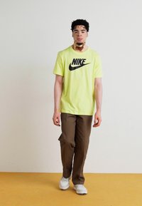 Camiseta de Nike en amarillo neón con logo negro, combinada con pantalones marrones y zapatillas blancas, sobre un fondo sencillo con suelo amarillo.