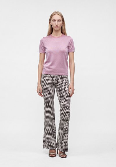 Femme aux longs cheveux blonds portant un top rose à manches courtes, un pantalon à motifs chevrons noir et blanc, et des sandales ouvertes, debout devant un fond uni.